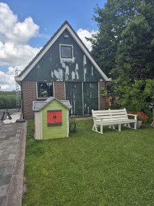 A small brick and wood Dutch style house with peeling green paint on the upper walls, a ladder on the side, and a white garden bench with a small green and red playhouse in the foreground.